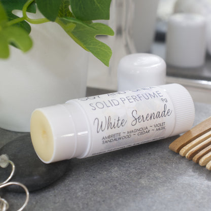 A white wind-up tube lays flat on a stone tray surrounded by accessories like earrings, watch and comb.  The lid has been removed and placed behind the tube revealing a beige balm.