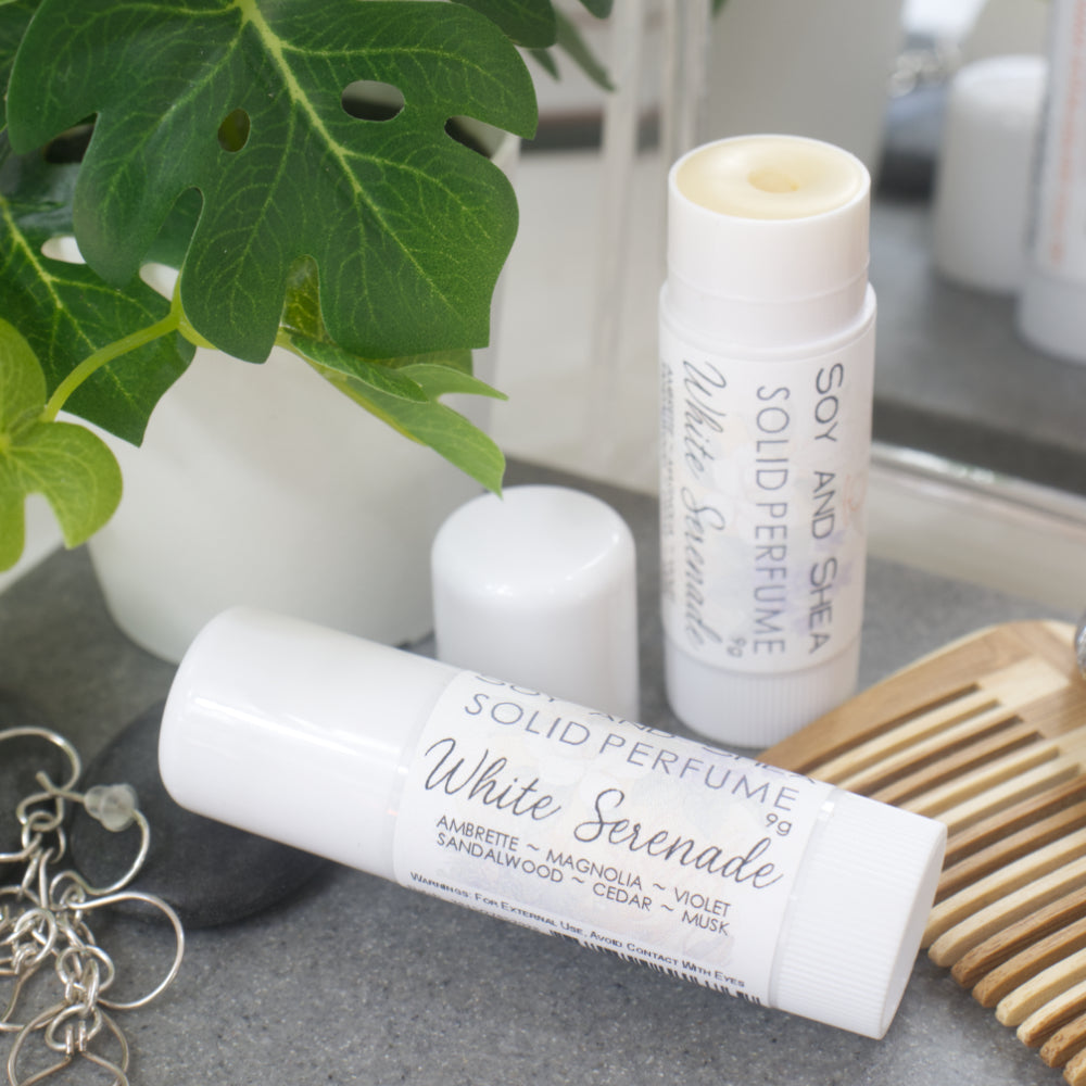 Two white tubes of Solid Perfume are displayed on a grey stone tray along with silver earrings, watch and greenery.   One tube lays flat  displaying the fragrance notes while another tube stands upright behin it with the lid removed and to the leftrevealing a beige balm