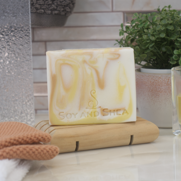 A bar of soap with a white and yellow marbled design, resting on a wooden soap dish with a plant in the background.
