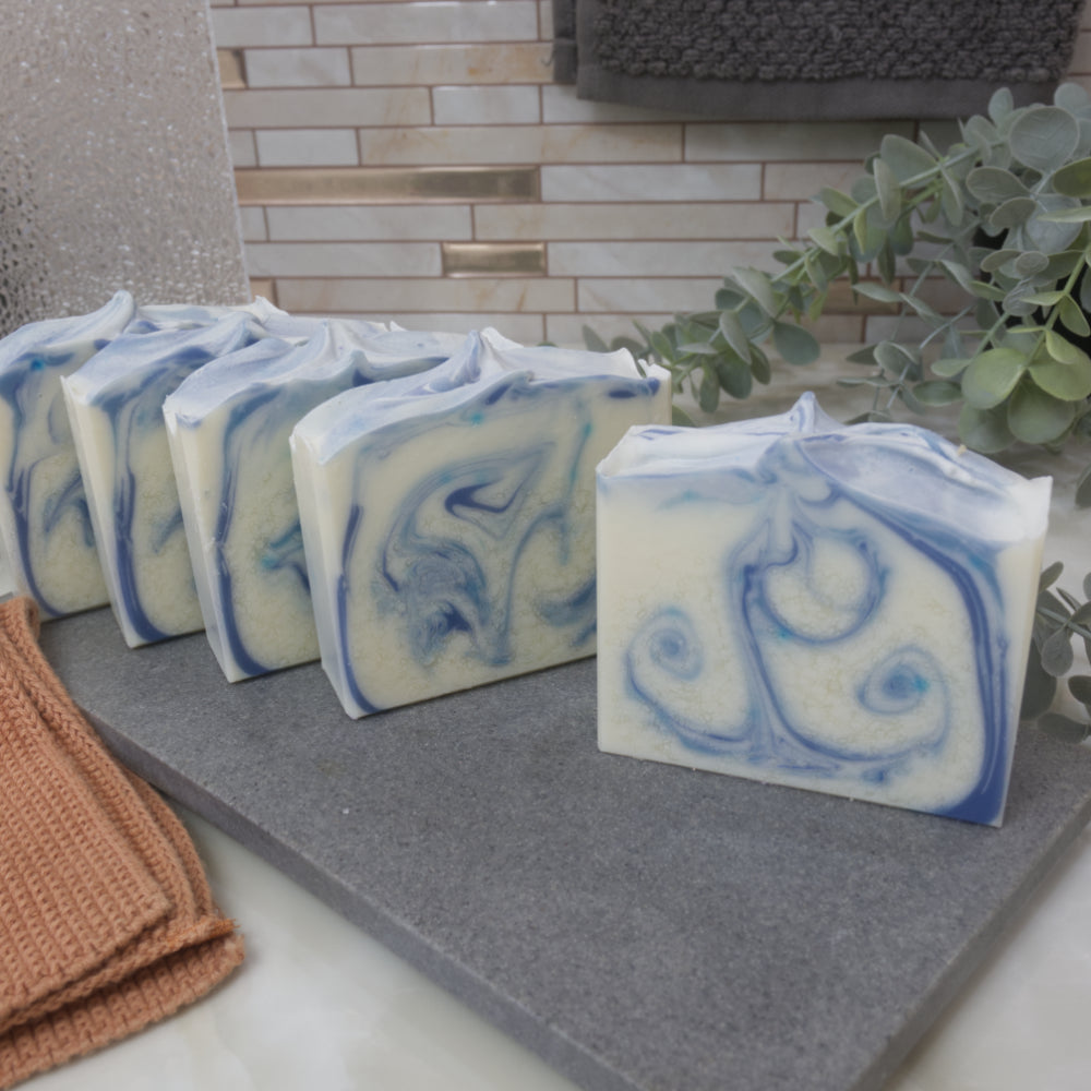 Five bars of soap sit one in front of the other off set enough to see the design on the edge of each soap.  The soaps have a main colour of white with a light and dark blue swirl.   These all sit on stone tray on a bathroom counter top with greenery and towels