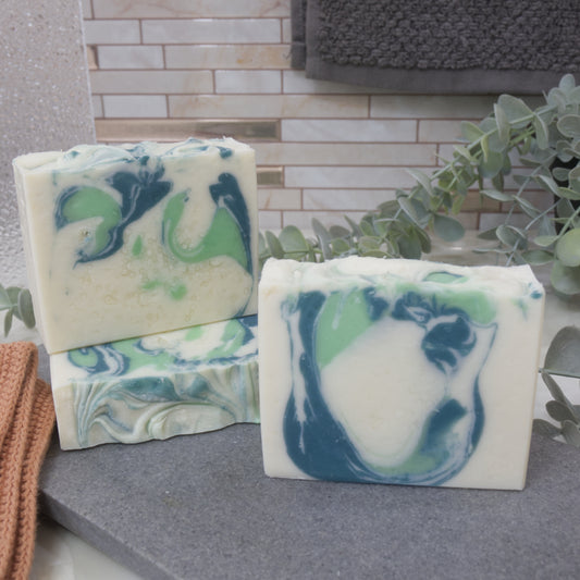 Three bars of soap on a gray surface with a tiled wall and plant in the background.   One soap is lying down with another standing upright on it.  In front is another soap bar.  All have a white base colour and feature various siwirl designs in a light and dark green
