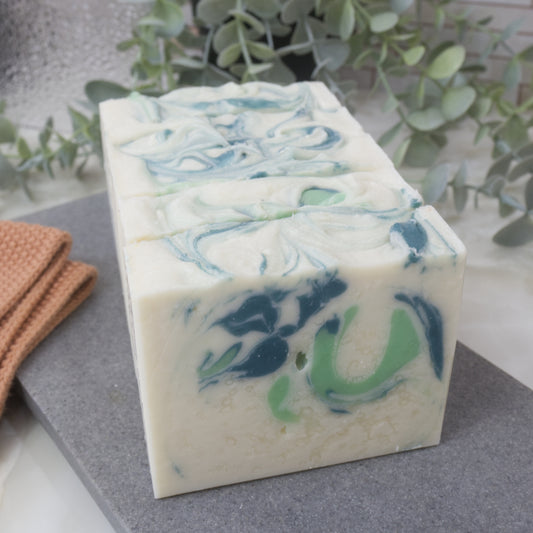 Five bars of soap sit one in front of the other with only the front bar showing the design.   The soap has a white main colour wiht swirls of Light and dark green.   These soaps sit on a grey stone tray surrounded by greenery and tiled background