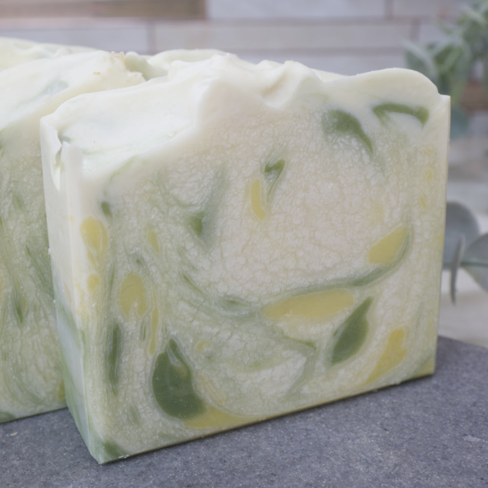 A close up of a soap bar that is white with a cracked effect through it.  There are splashes of dark green and yellow through the soap.  This sits on a grey stone bench with blurred background