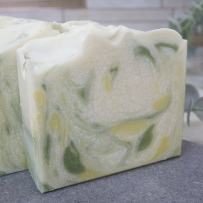 A close up of a soap bar that is white with a cracked effect through it.  There are splashes of dark green and yellow through the soap.  This sits on a grey stone bench with blurred background