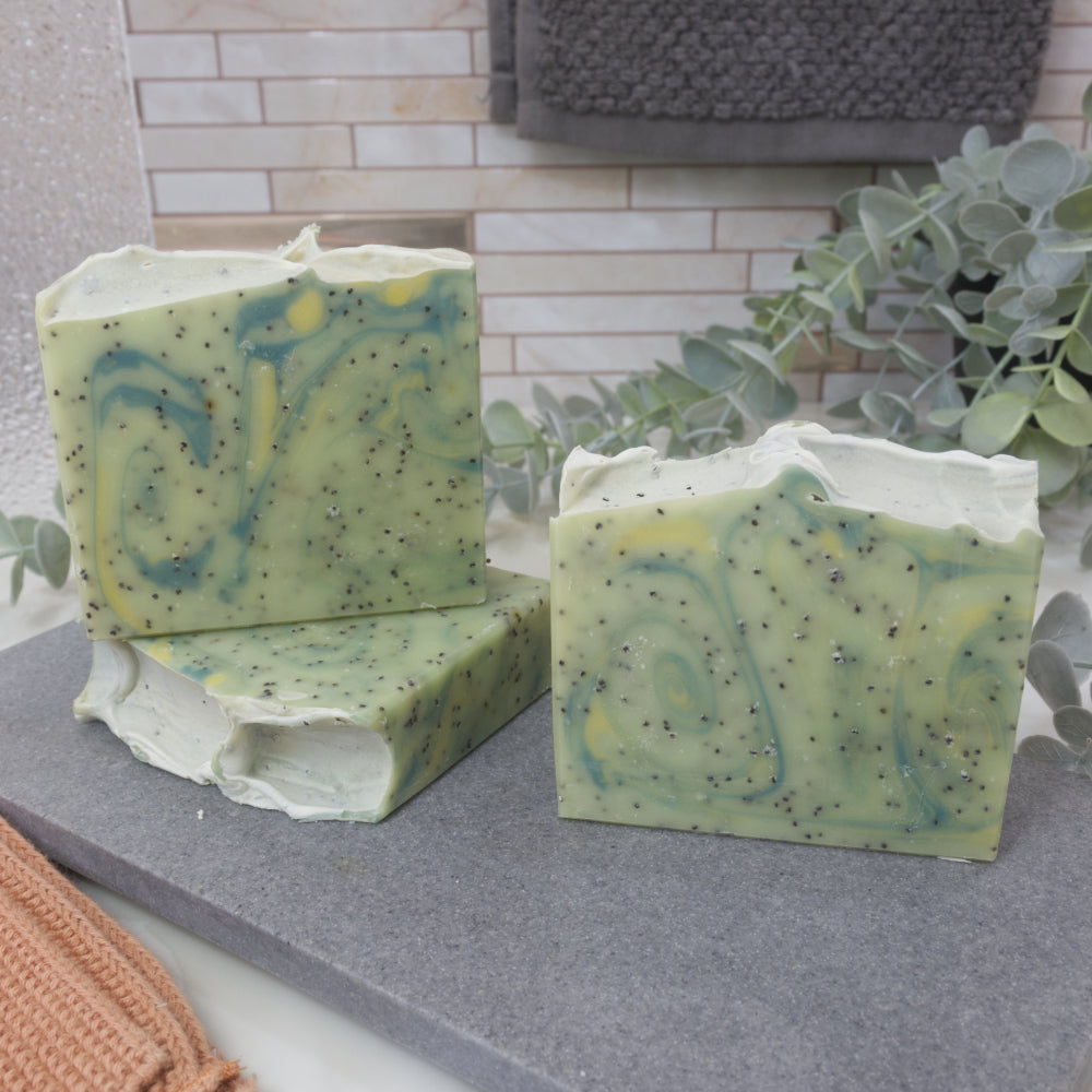 Three soap bars of pale green with dark green and yellow swirls sit on a gray stone tray with a tiled wall and plant in the background.