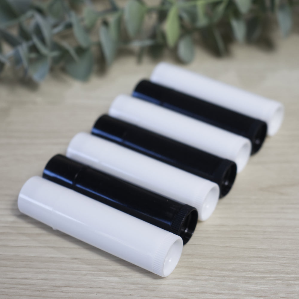 Seven lip balm tubes lie on a wooden surface with greenery in the background.  The tubes alternate in colour starting with white, followed by a black