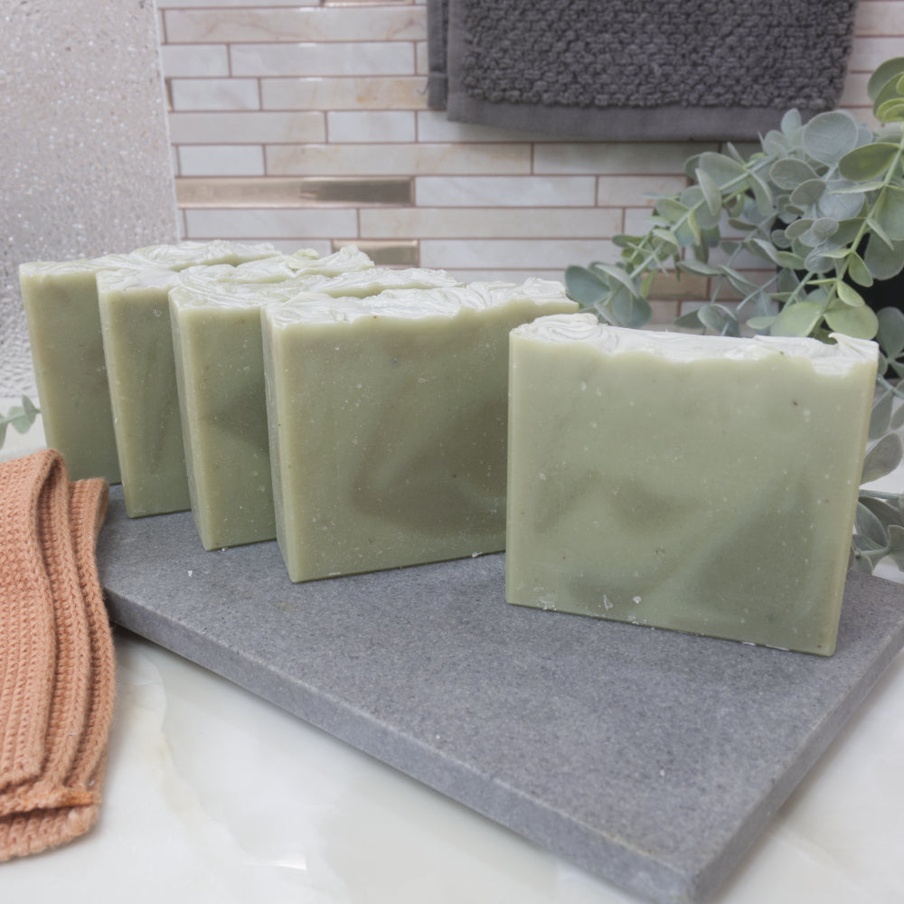 Five pale green soap bars  wiht dark green swirls on a gray stone surface with a towel and plant in the background
