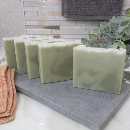 Five pale green soap bars  wiht dark green swirls on a gray stone surface with a towel and plant in the background