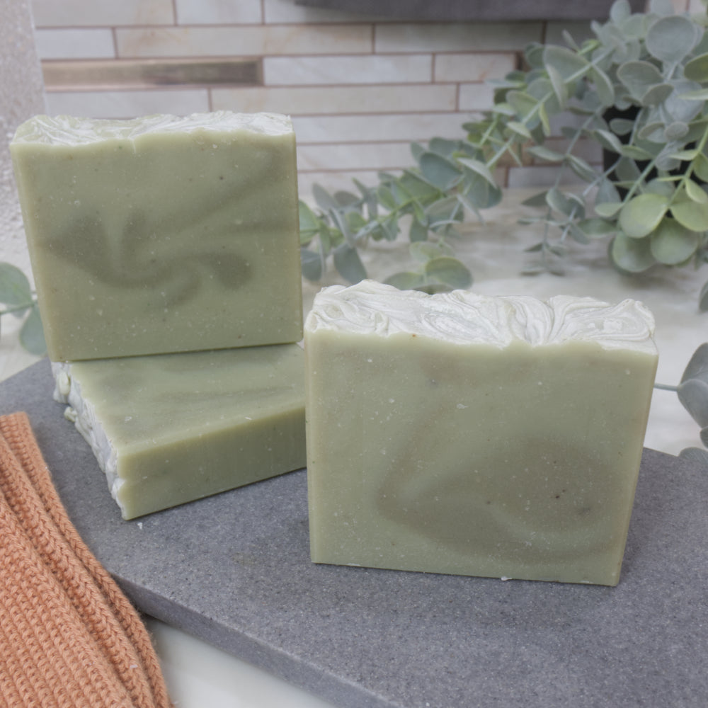 Three pale green bars of soap  with dark green swirls on a gray surface with a plant in the background