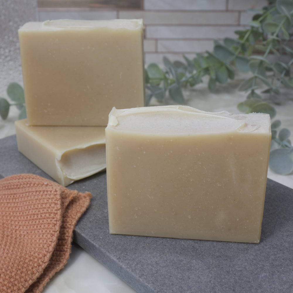 Three bars of cream soap on a gray surface with a brown towel and green leaves in the background