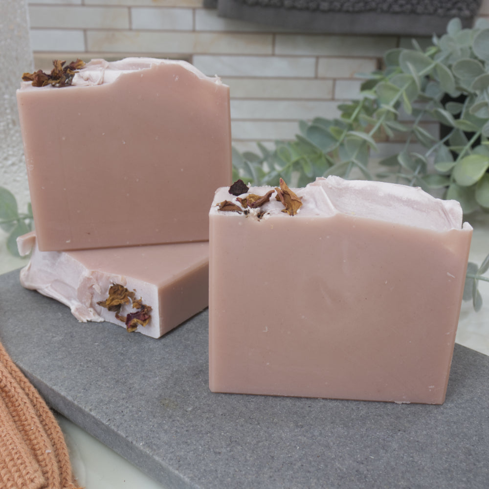 A selection of dusky pink soaps with rose petals on top sit on a grey stone surface against a tile background with greenery
