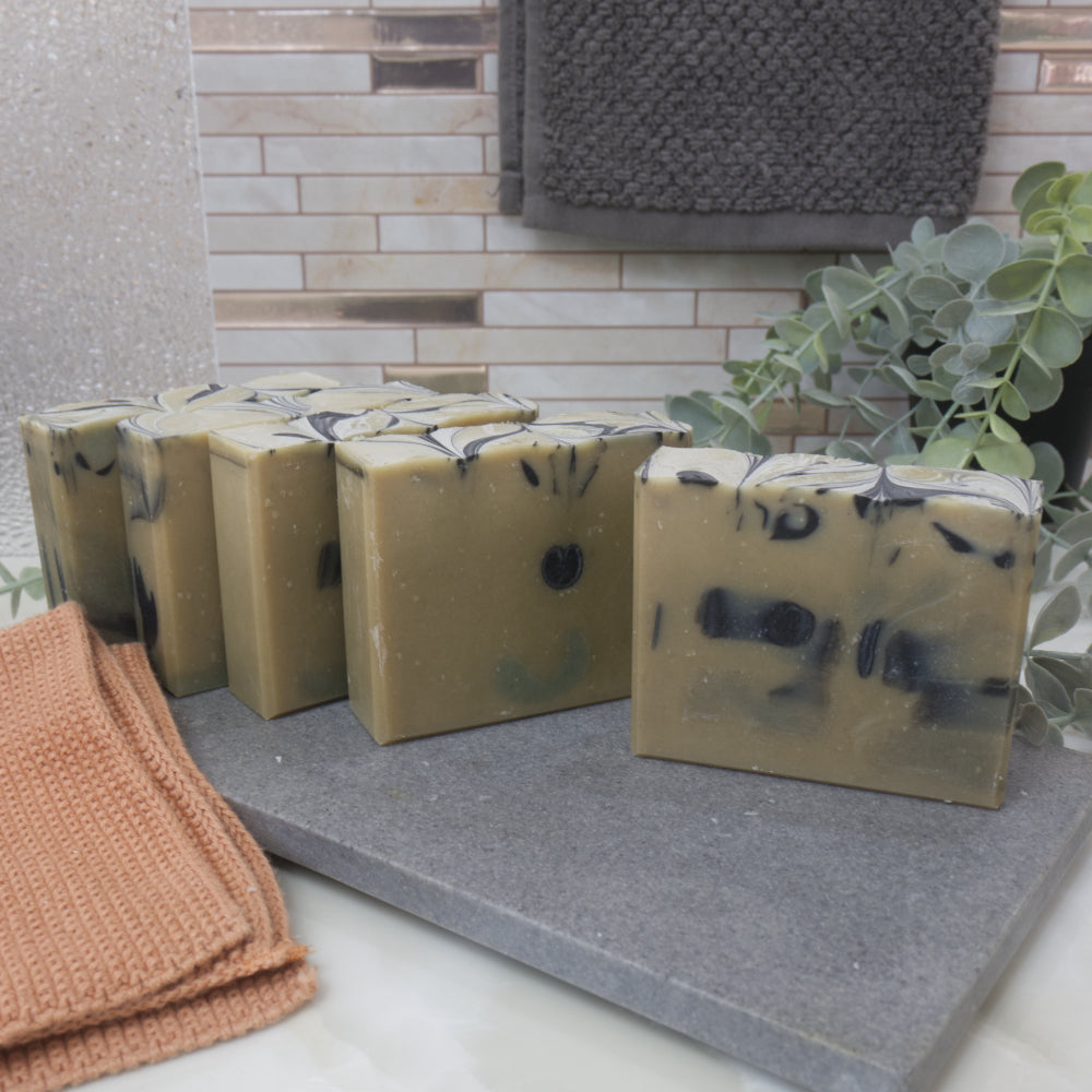 A selection of green soaps with black swirls are lined up on a stone surface, with a towel and plant in the background.