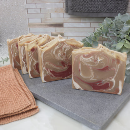 Five bars of soap bars are lined up on a gray stone tray with a textured wall and plant in the background.  The soaps are placed one in front of the other and slightly offset so see a peack of the design.  The front two soaps show soaps that are light brown in colour with decorative swirls in red, brown and white.