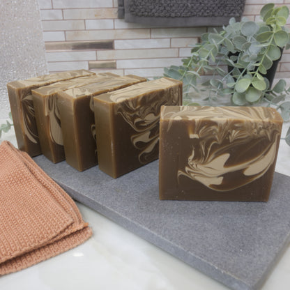 Brown soap bars with cream swirl patterns on a gray surface, with a towel and plant in the background.