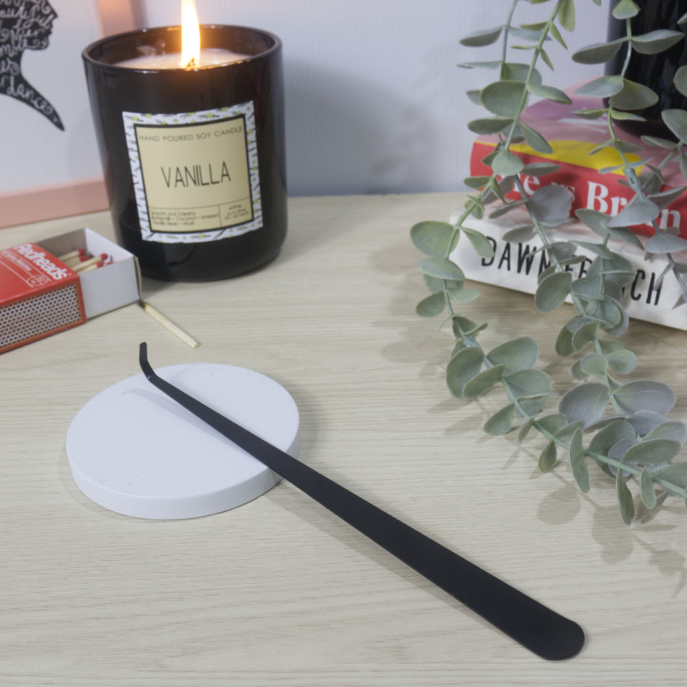 A long narrow black metal tool with a hooked end rests on a white candle lid. In the background is a lit Vanilla candle in a black jar along with an open box of matches, a stack of books and greenery