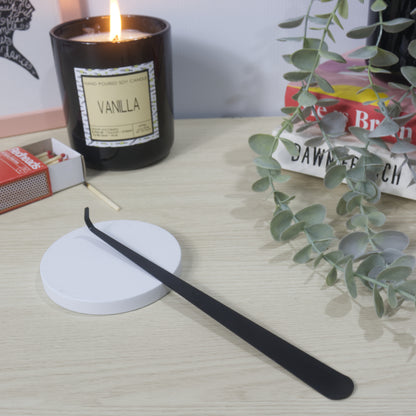 A long narrow black metal tool with a hooked end rests on a white candle lid. In the background is a lit Vanilla candle in a black jar along with an open box of matches, a stack of books and greenery