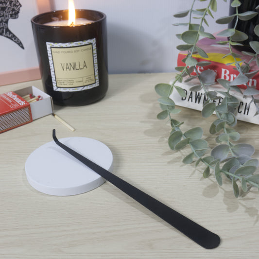 A long narrow black metal tool with a hooked end rests on a white candle lid. In the background is a lit Vanilla candle in a black jar along with an open box of matches, a stack of books and greenery
