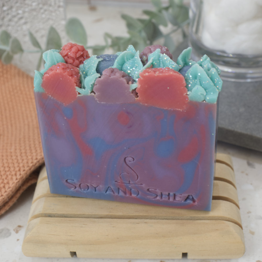 A bar of soap sits on a wooden soap dish with a blurred background.  The soap has a marble design of purple, red and blue and features a decorative top of berries and leaves made from soap