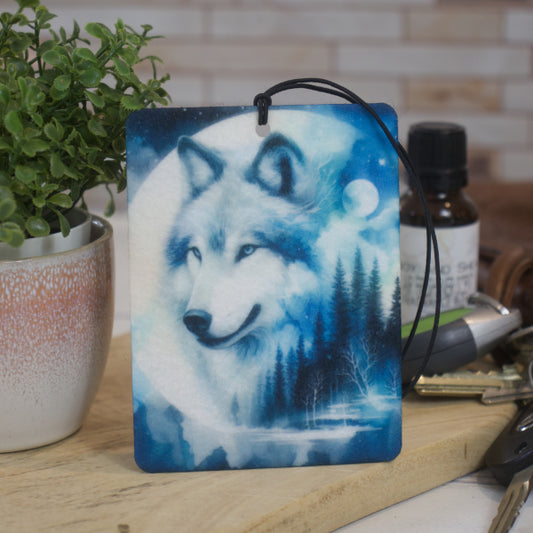 A car air freshener featuring an artistic depiction of a wolf with a moon and forest background rests against a pot plant next to a bottle of fragrance and a set of keys.