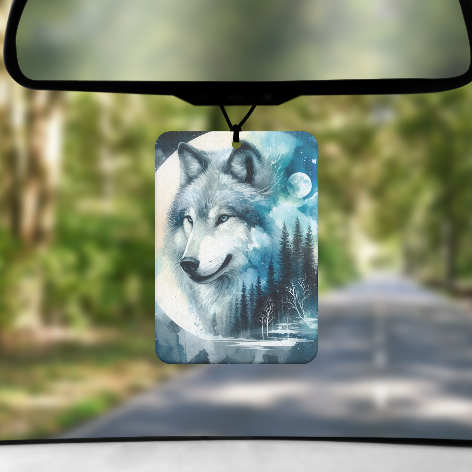 A mock up image showing a view through a car windscreen with a long tree lined road.  From the rearview mirror hangs an air freshener with an image of a wolfs head in a moon and the reflection of a lake and trees
