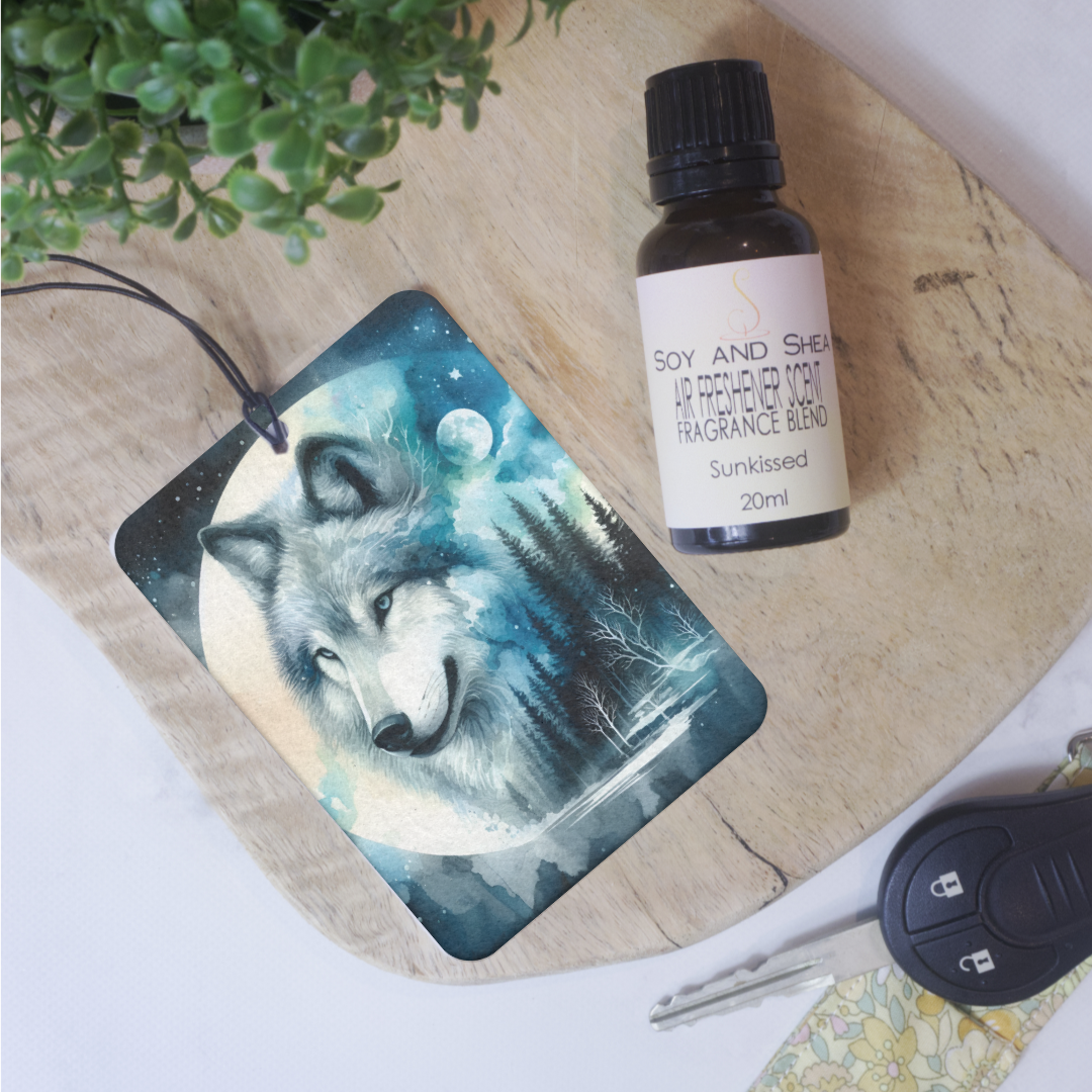 A car air freshener featuring an artistic depiction of a wolf with a moon and forest background, placed on a wooden surface.  Next to is a glass bottle of Air Freshener Scent  