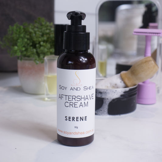 A bottle of Serene Aftershave Cream with a pump dispenser, placed on a bathroom countertop with a plant and other bathroom accessories in the background including shaving soap, brush and pink razor.