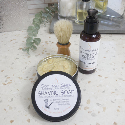 A black, round tin of shaving soap sits on a bathroom counter top with the lid off and resting on the side revealing a cream soap inside.  Behind stands a shaving brush with wooden handle and next to that is a amber bottle of Aftershave cream with lotion pump lid.