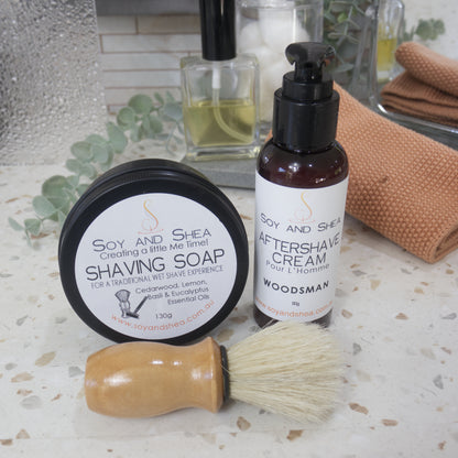 A black, round tin of shaving soap sits on its side with the lid facing the camera.  In front, laying flat is a shaving brush with wooden handle.  Next to the Shaving soap is a bottle of Aftershave cream with a lotion pump lid.  Behind is a blurred background of a bathroom countertop