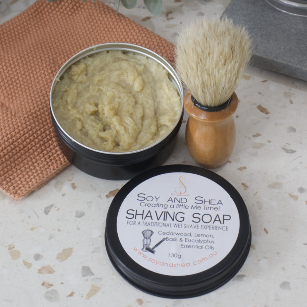 A black tin of fresh Shaving Soap sits on a bathroom counter top with the lid sitting to one side.  The Soap is a blue cream/yellow colour.  A shaving brush stands next to the tin.