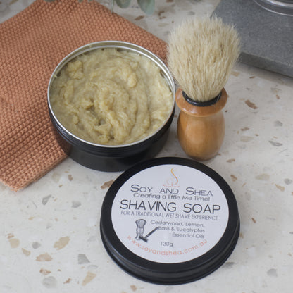 A black tin of fresh Shaving Soap sits on a bathroom counter top with the lid sitting to one side.  The Soap is a blue cream/yellow colour.  A shaving brush stands next to the tin.