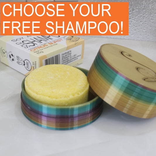 A Yellow shampoo bar sits in a colorful container with a box of soap in the background and text 'CHOOSE YOUR FREE SHAMPOO!' at the top.
