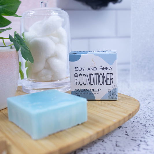 A wooden tray sits on a bathroom counter with a potted plant and jar of cotton balls against a blurred tiled background. Towards the front of the tray stands a white box with blue abstract pattern with text SOLID CONDITIONER and next to it stands a square reen-blue bar of conditioner.