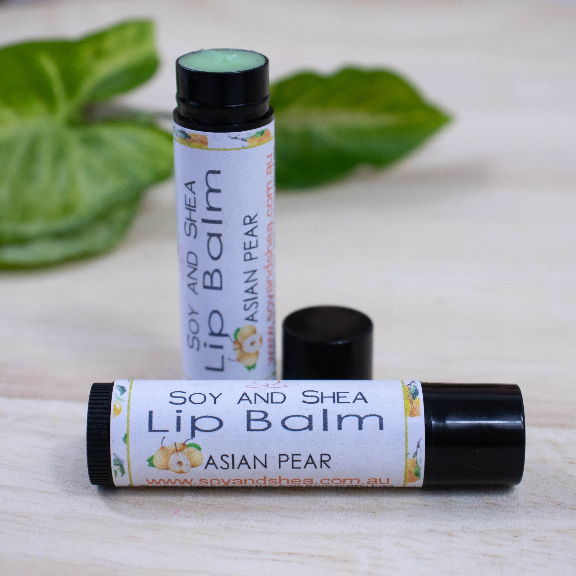 Two black tubes of lip balm displayed on a wooden surface with greenery blurred in the background.  One tube lies flat across the surface displaying the flavour while the other tube stands upright behind it with the cap removed and placed to the side revealing a green balm.