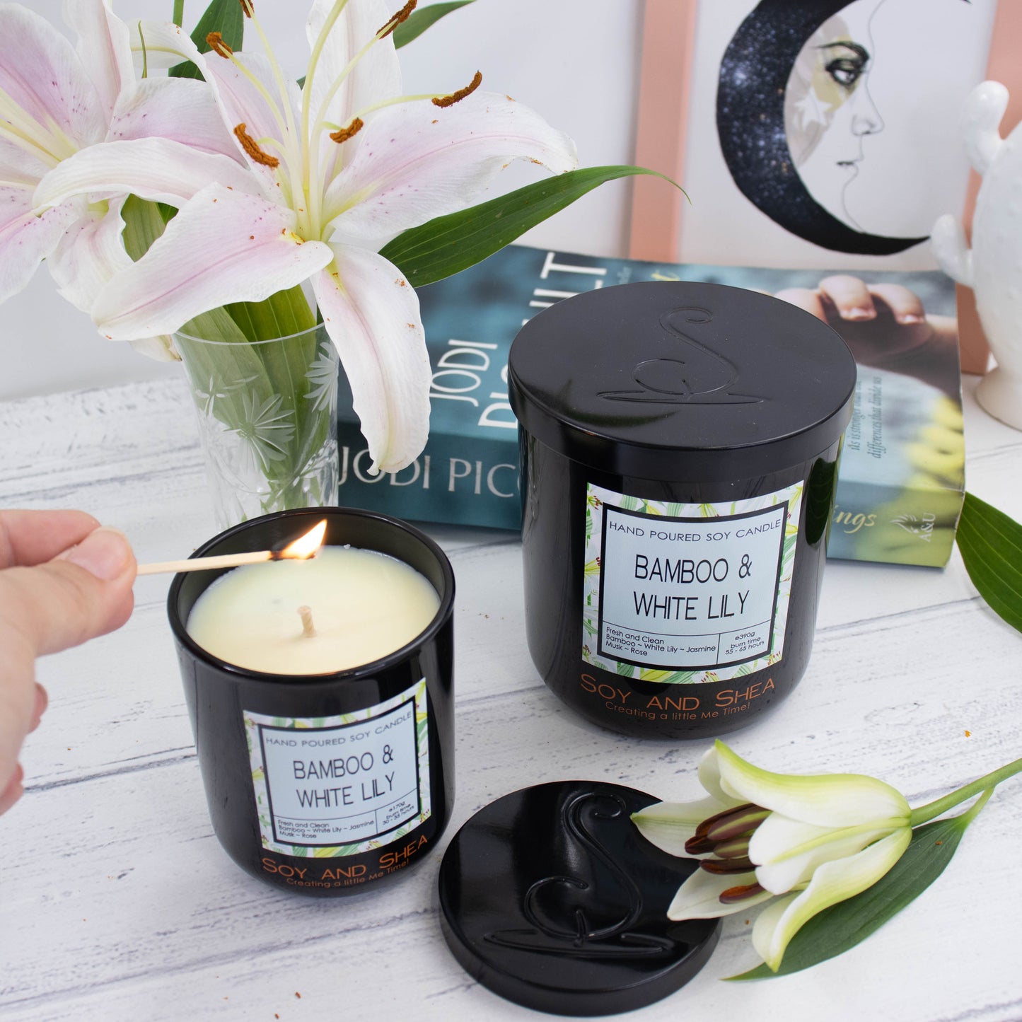 A large black glass candle with decorative product label and black metal lid stands on a white wooden surface in front of a book, picture frame and vase of white lillies.   To the front is a smaller candle with its lid removed and placed on the side to reveal a pale green wax.  A hand reaches in with a lit match to light the candle.  The lid of the candles show a debossed logomark.