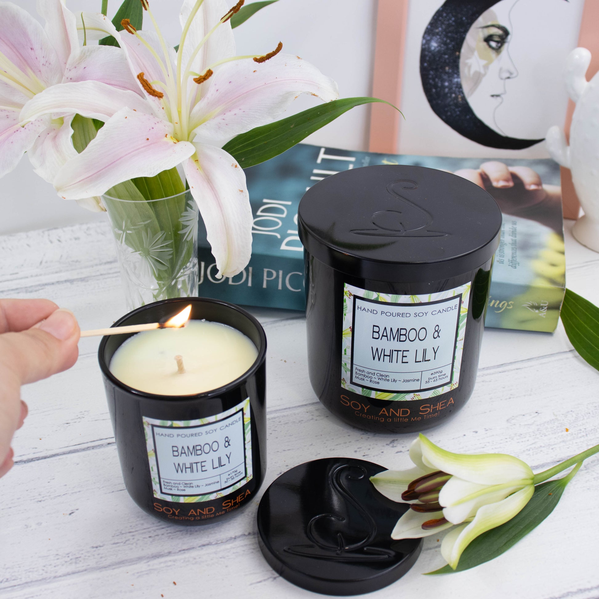 A large black glass candle with decorative product label and black metal lid stands on a white wooden surface in front of a book, picture frame and vase of white lillies.   To the front is a smaller candle with its lid removed and placed on the side to reveal a pale green wax.  A hand reaches in with a lit match to light the candle.  The lid of the candles show a debossed logomark.