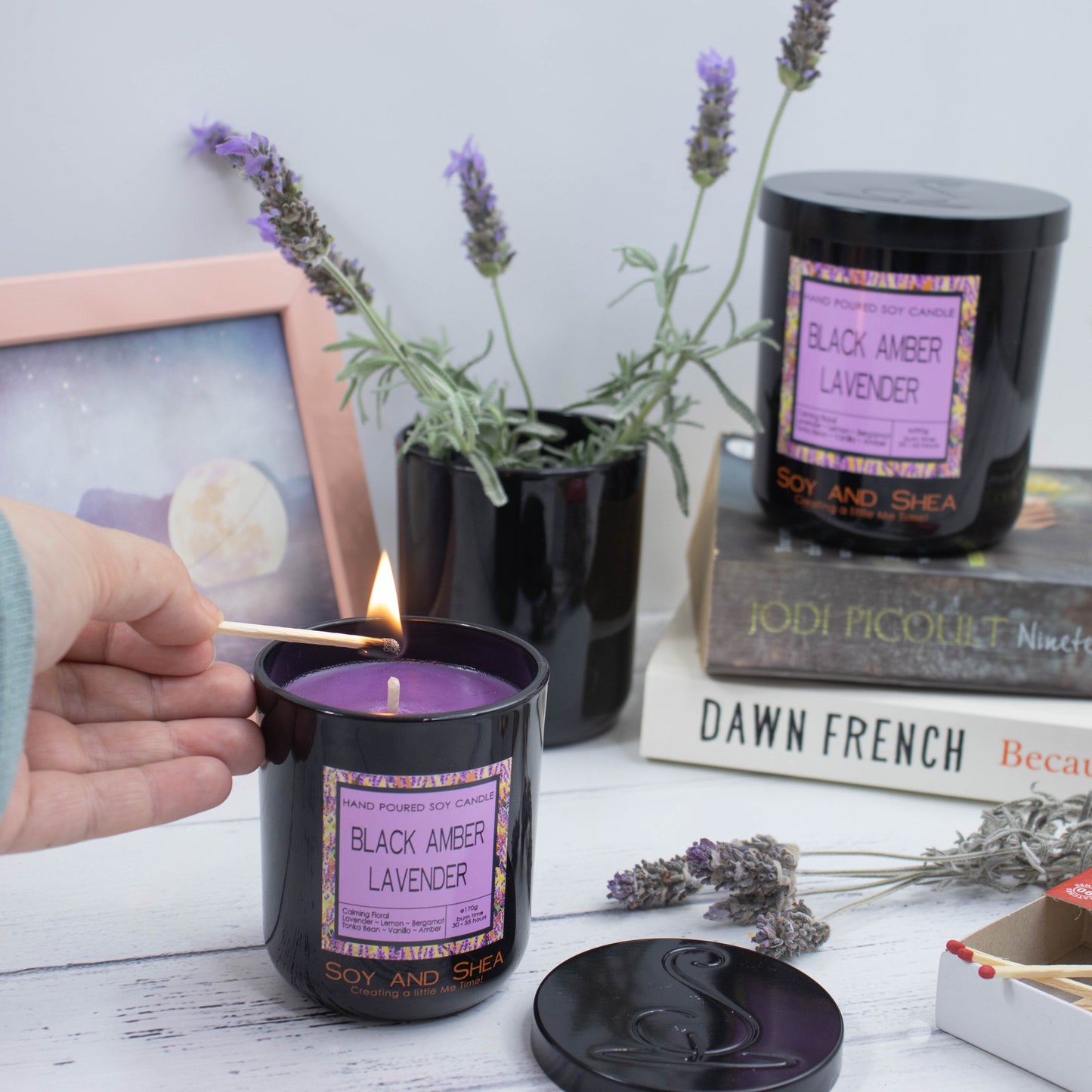 A black candle jar filled with purple wax is about to be lit with a match.  To the right is the lid which has an engraving of the logo.  In the background is a photo frame and  and to its right is a pot of fresh lavender.   To the right is a stack of books with a larger candle sitting on top.