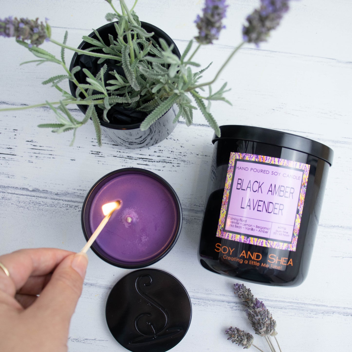 A selection of candles in black jars are displayed on a wooden bench.  A large candle lays on its side with the label facing up while to the left, a small jar filled with purple wax is being lit with a match.  A pot of Lavender is sits in the corner