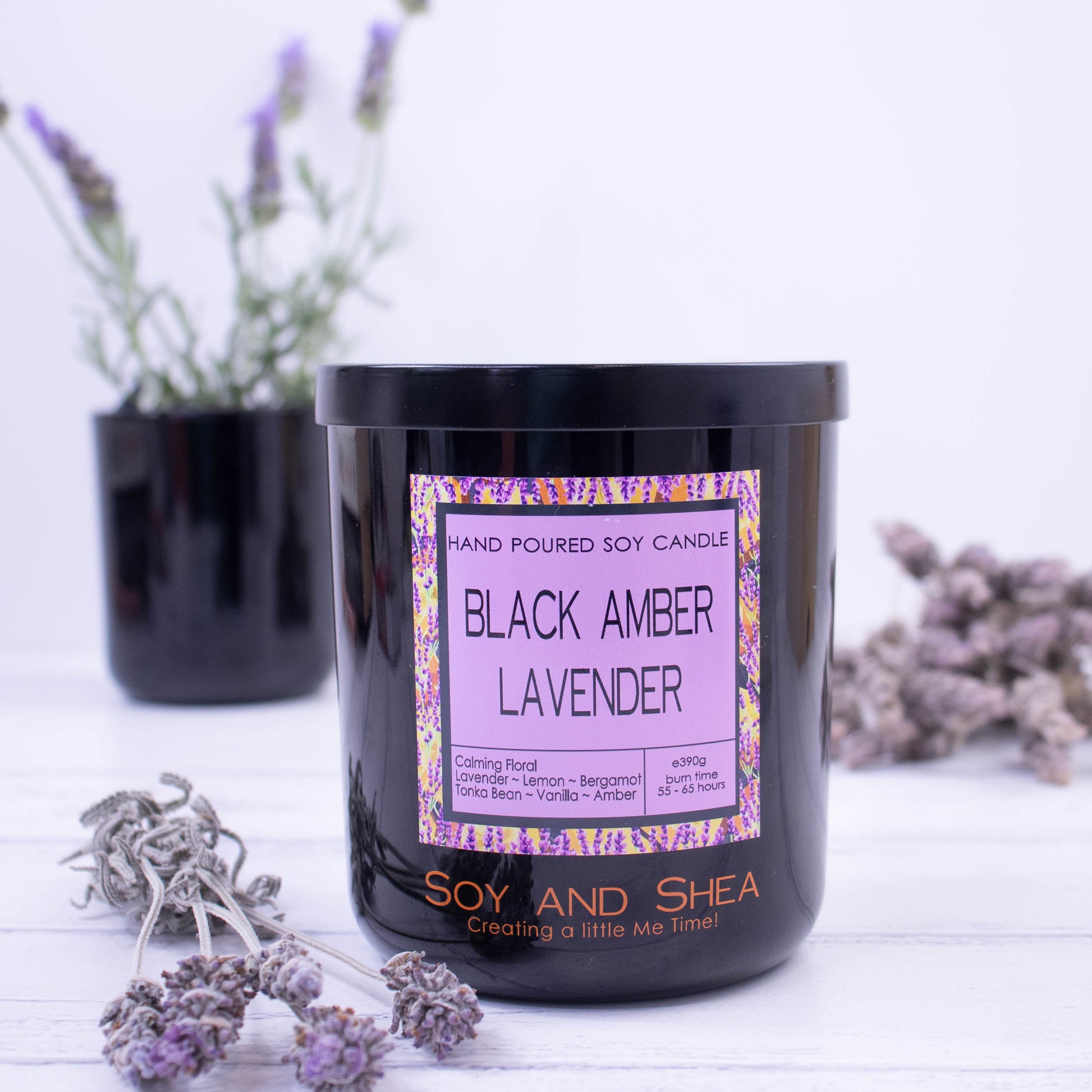 A large black glass candle jar with fragrance label sits to the front of the picture.  There is a black metal lid with an etching of the logo on top.  Surrounding the candle is some dried lavender with a pot of fresh lavender growing in the background