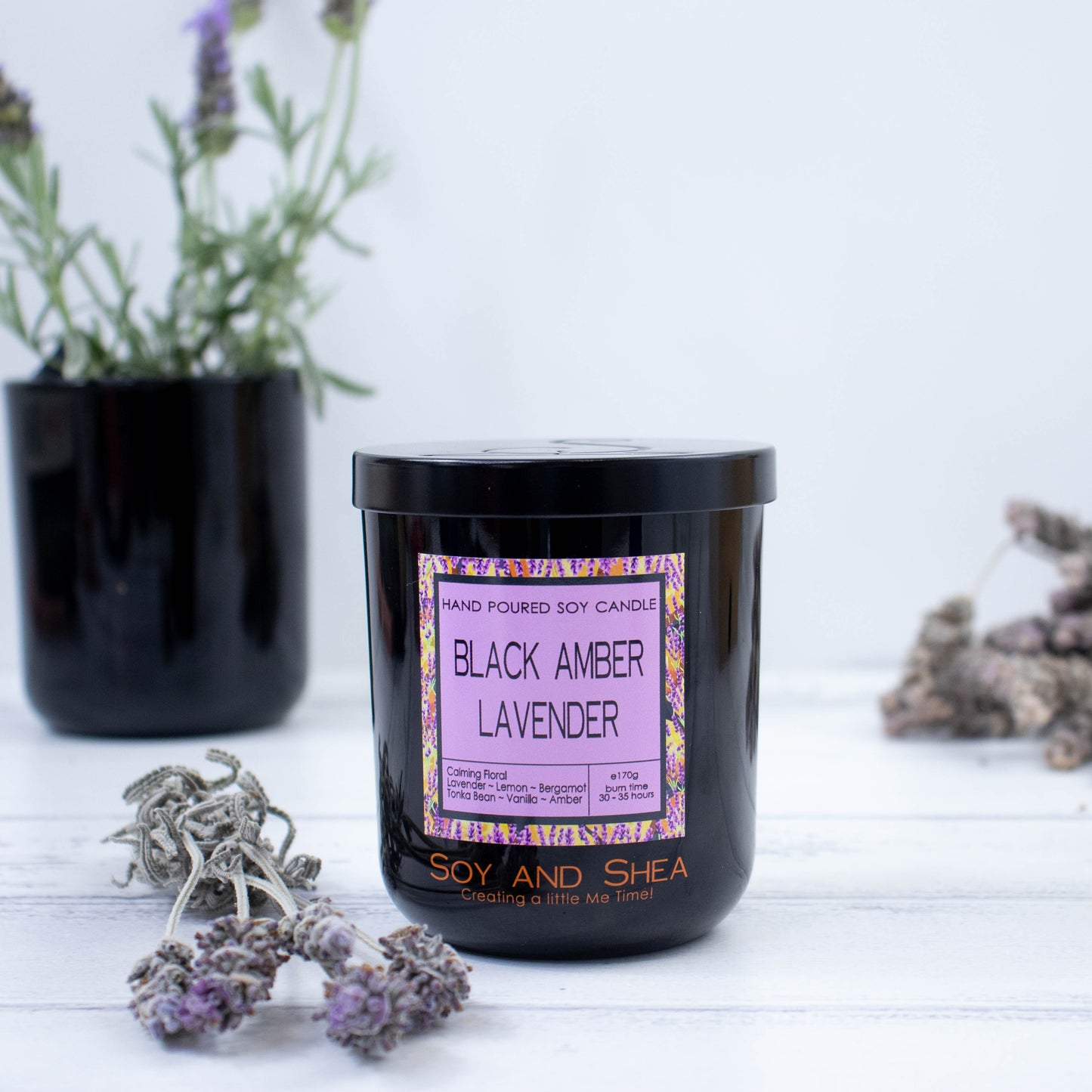 A black glass candle jar with fragrance label sits to the front of the picture.  There is a black metal lid with an etching of the logo on top.  Surrounding the candle is some dried lavender with a pot of fresh lavender growing in the background