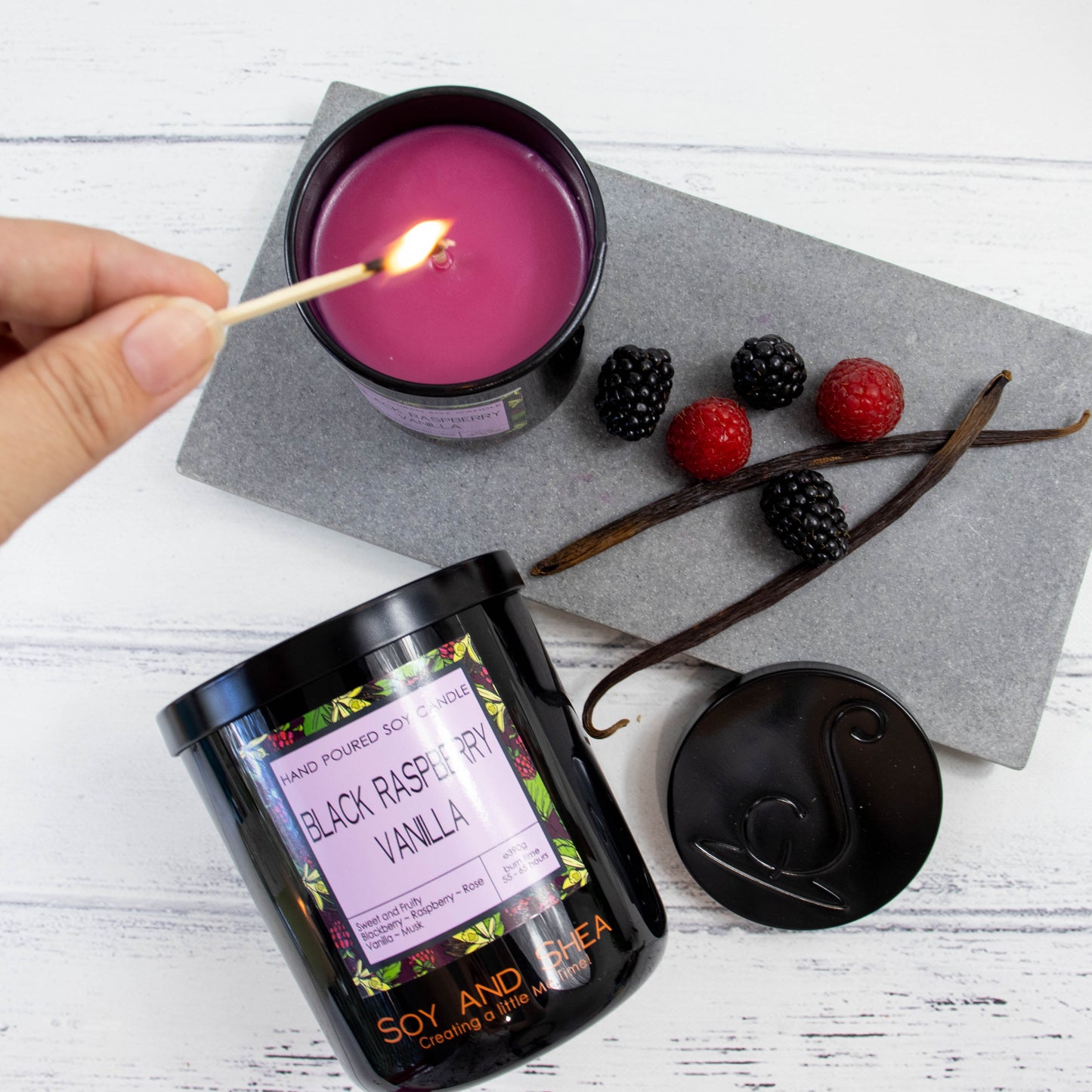 A flat lay photo showing a large black jar lying on its side on a white wooden surface toward the bottom of the image.  Above it is a grey stone tray with a selection of red and black berries and two dried vanilla pods.  A small candle stands on the tray with a hand reaching in with a lit match to light the magenta wax.  The black metal lid of the candle rests against the edge of the tray showing the debossed image of the logomark.