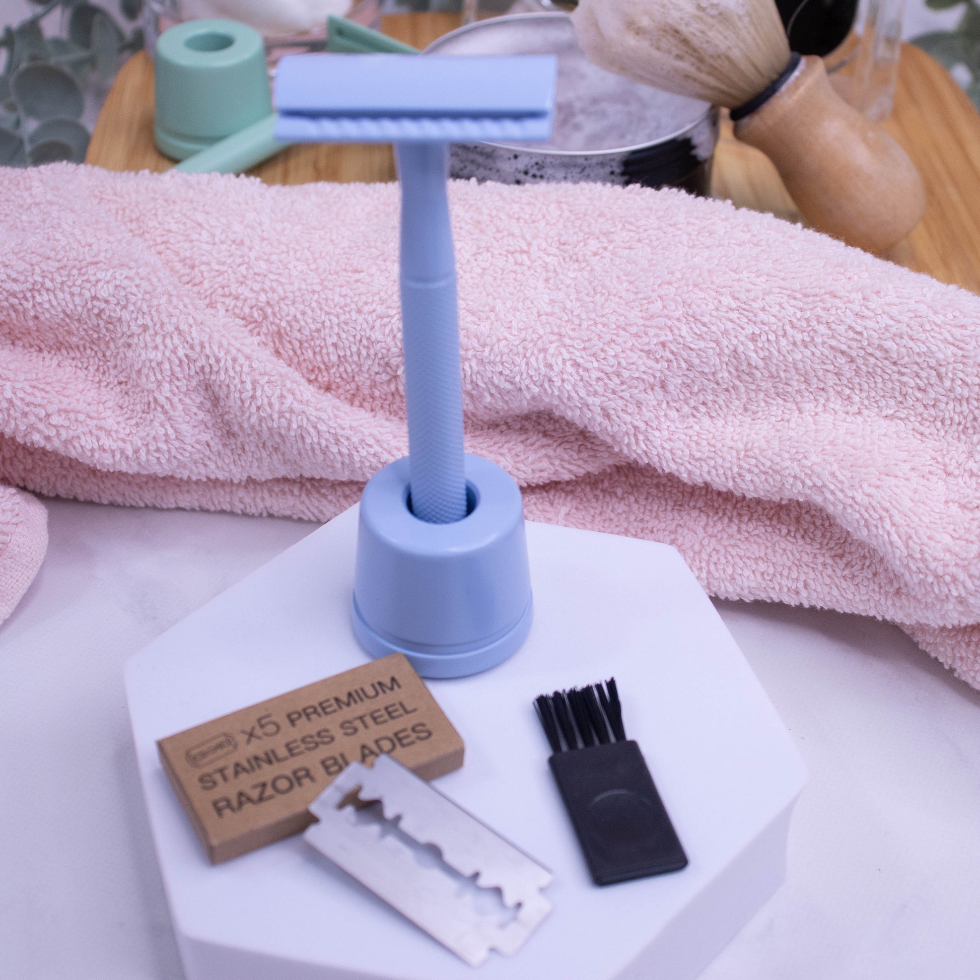 A pale blue safety razor stands upright in its matching holder with a pack of five stainless steel blades and small brush in front on a white foam stand.  Behind is a pink towel and a tin of Shaving soap and a brush resting on a wooden tray.