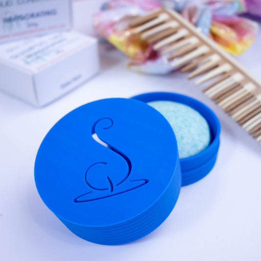A bright blue cylindrical pot with the Soy and Shea logomark cut out in the top, sits on a white surface.  The lid has been removed and is resting on the side of the base which holds a solid shampoo bar.  In the background stands the shampoo packaging next to a wooden comb and colourful scrunchie with shiny gold dots.