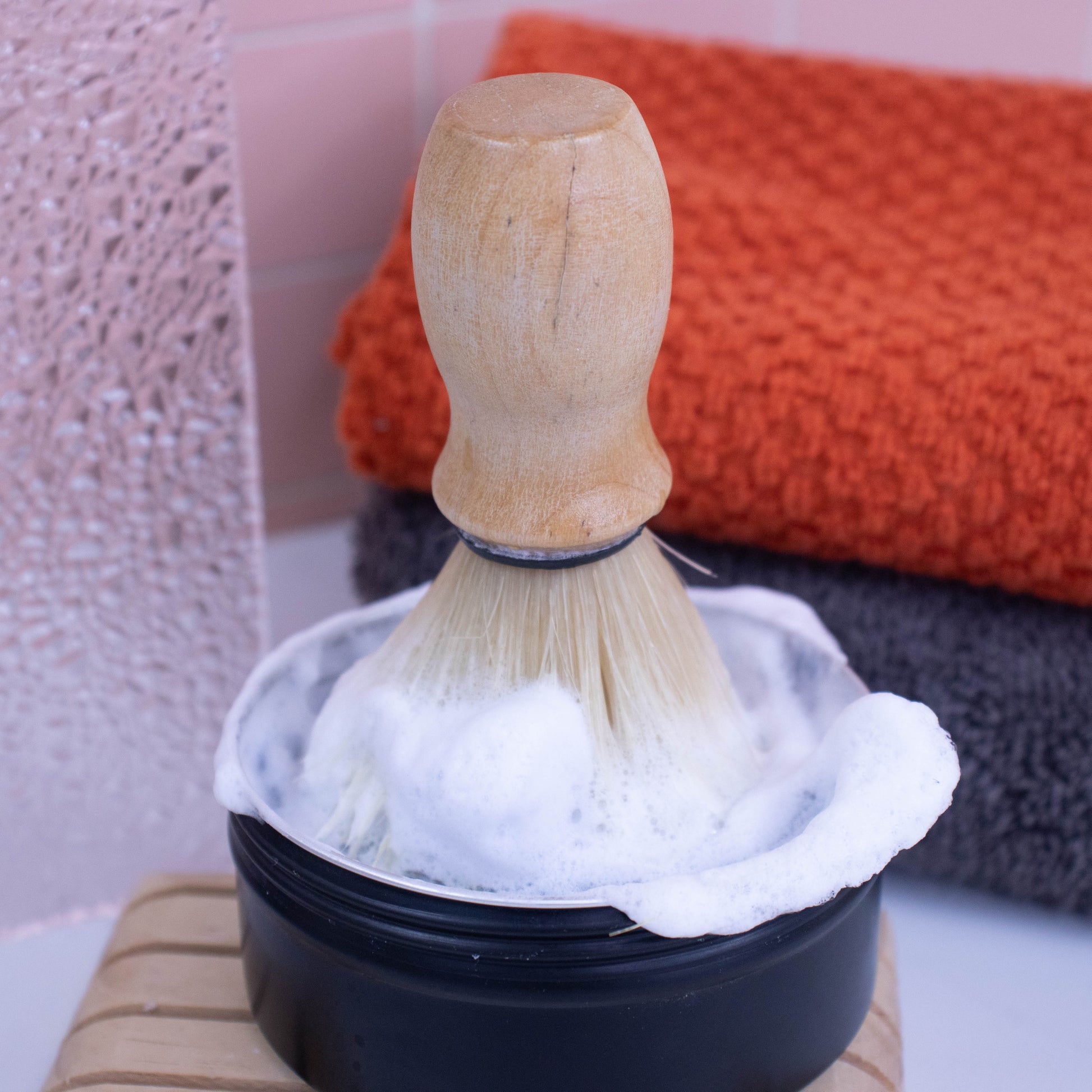 A shaving brush with wooden handled stands upright with its bristles in a black tin of foam from soap.  Behind is a stack of grey and orange towels.