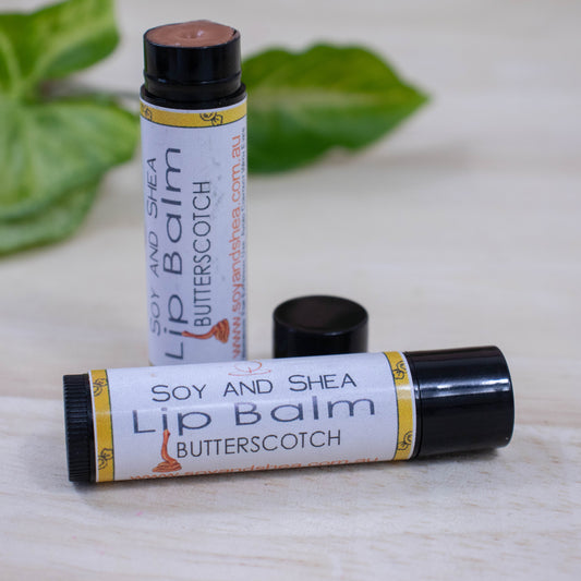 Two black tubes of lip balm displayed on a wooden surface with greenery blurred in the background.  One tube lies flat across the surface displaying the flavour while the other tube stands upright behind it with the cap removed and placed to the side revealing a brown balm.