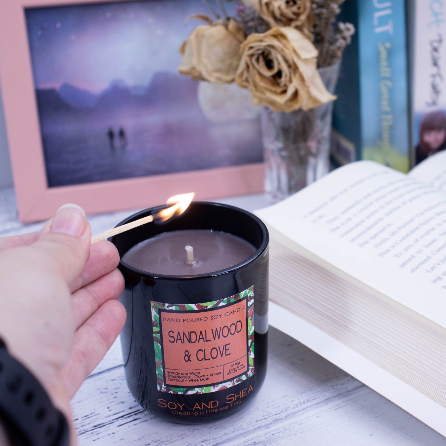 A black glass candle filled with brown wax is about to be lit with a match. There is an open book to the side of the candle with other books in the background along with a vase of dried flowers and a picture in a pale pink frame