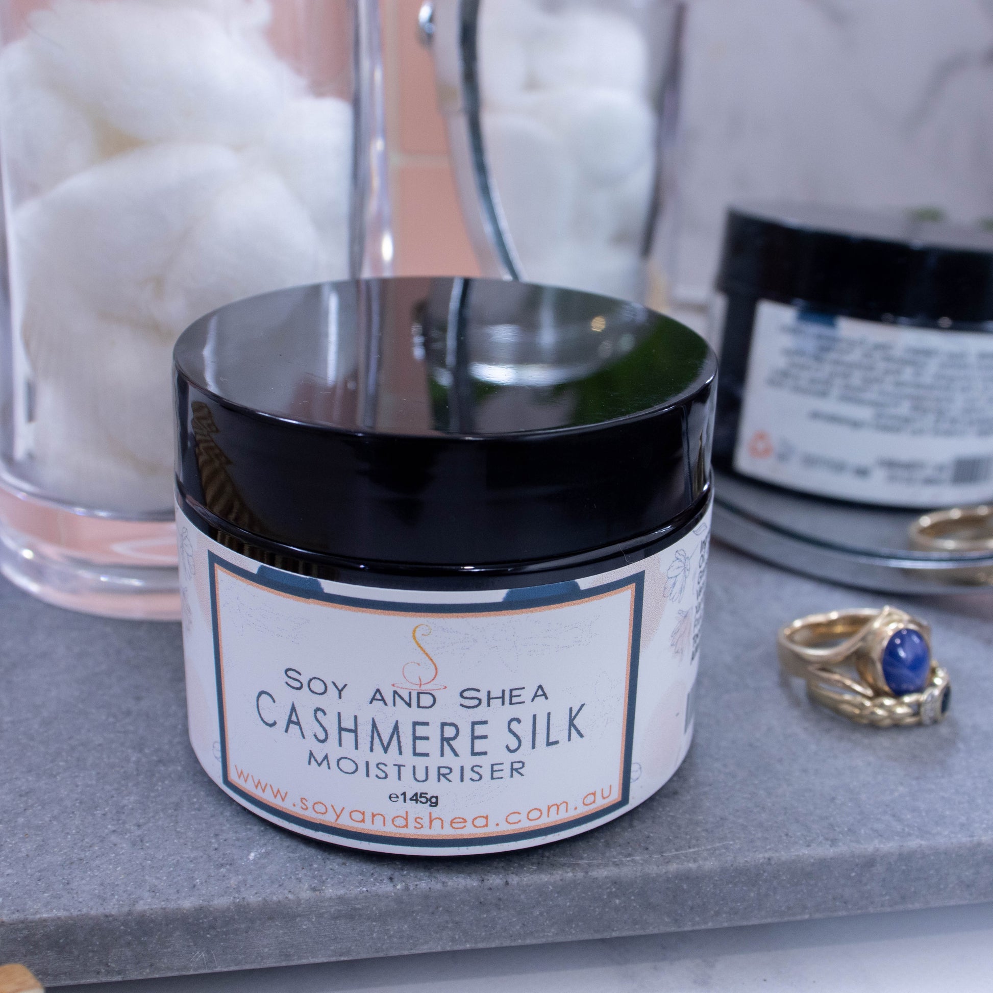 A jar with black lid stands on a grey stone surface surrounded by a jar of white cotton balls, gold rings and a mirror reflecting the back of the jar.  The jar has a white label listing the scent and has hints of blue and orange 