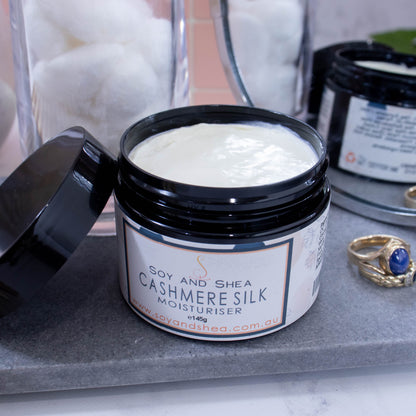An open jar of moisturiser labelled CASHMERE SILK, sits on a grey stone surface next to jar of white cotton balls, some gold rings and a mirror reflecting the back of the jar.   The jar is black and has a black lid resting on the side revealing an off-white cream inside.