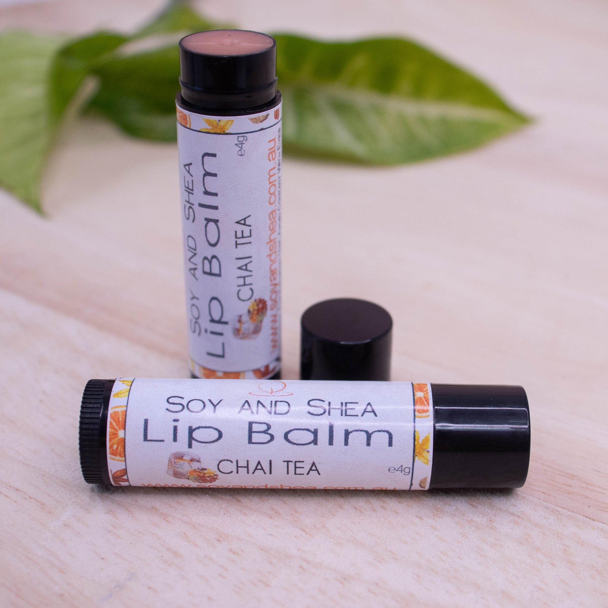 Two black tubes of lip balm displayed on a wooden surface with greenery blurred in the background.  One tube lies flat across the surface displaying the flavour while the other tube stands upright behind it with the cap removed and placed to the side revealing a brown coloured balm.