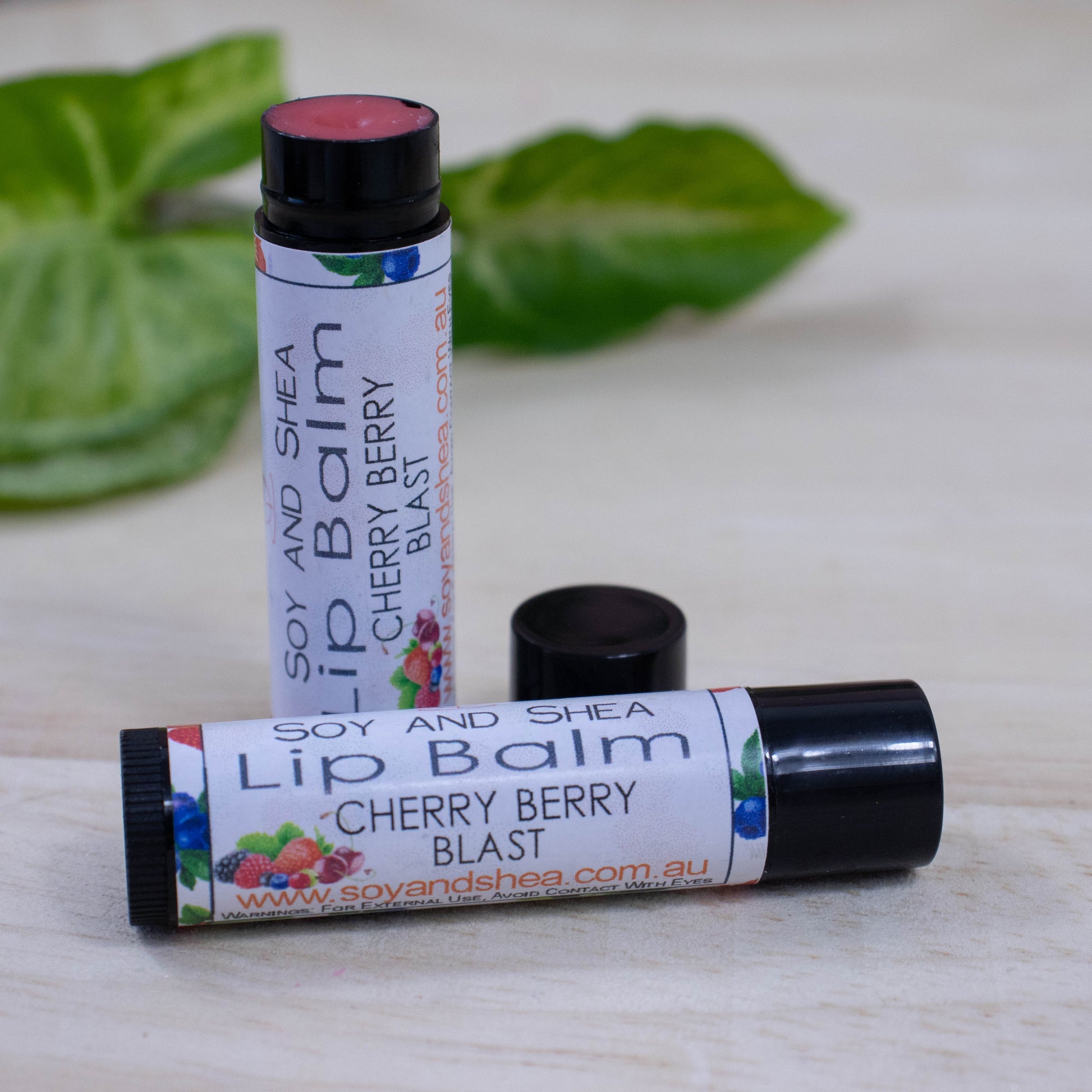 Two black tubes of lip balm displayed on a wooden surface with greenery blurred in the background.  One tube lies flat across the surface displaying the flavour while the other tube stands upright behind it with the cap removed and placed to the side revealing a red balm.