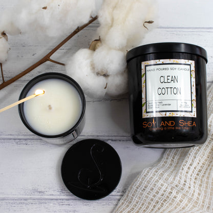 A flat lay photo showing a large black jar lying on its side on a white wooden surface.  To the left of the jar is a smaller candle standing upright, filled with white wax  and a hand reaches in with a lit match. Below is a round black metal lid with the logomark debossed in the centre.  The candles are surrounded by natural cotton bolls and a cream cottom cloth.